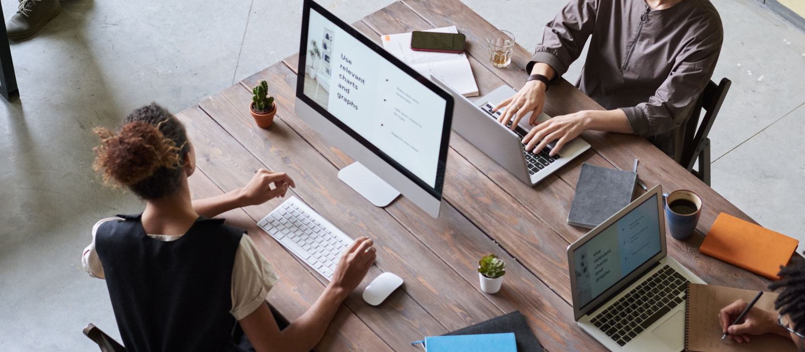 A project team working on laptops and a desktop at a shared wooden desk, seen from above
