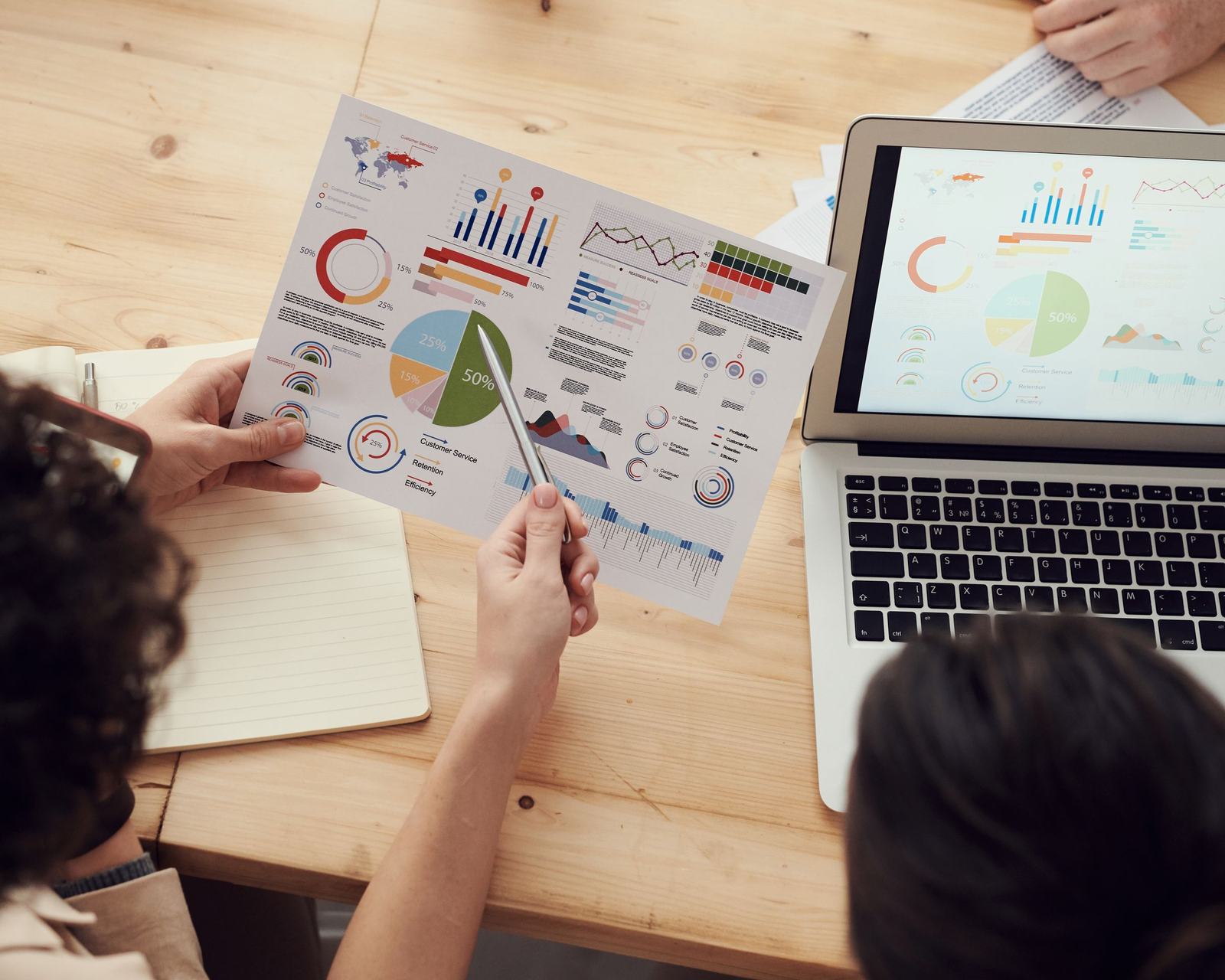 A consulting team reviewing printed charts and a laptop at a wooden table, seen from above, with margin, expense and service-report cards from Odoo floating above