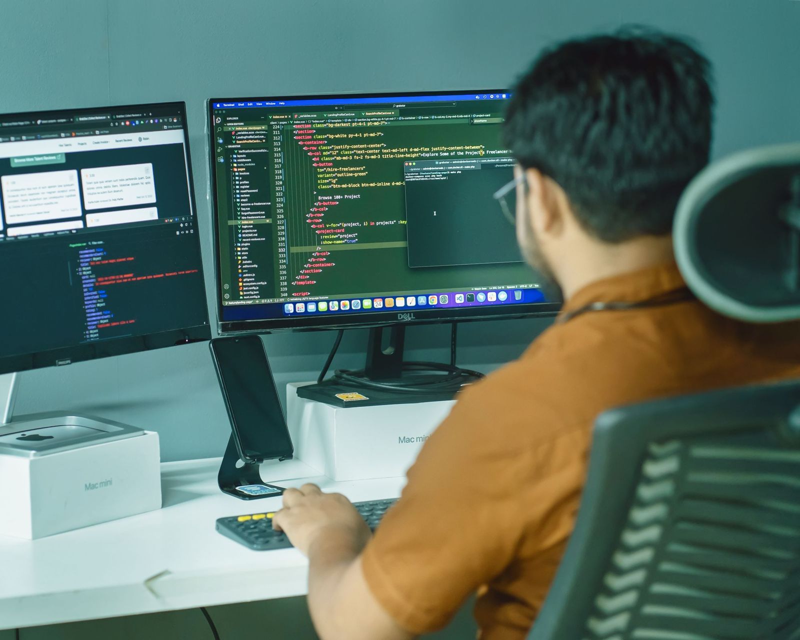 An engineer seen from behind at a desk with two monitors showing code and a log view