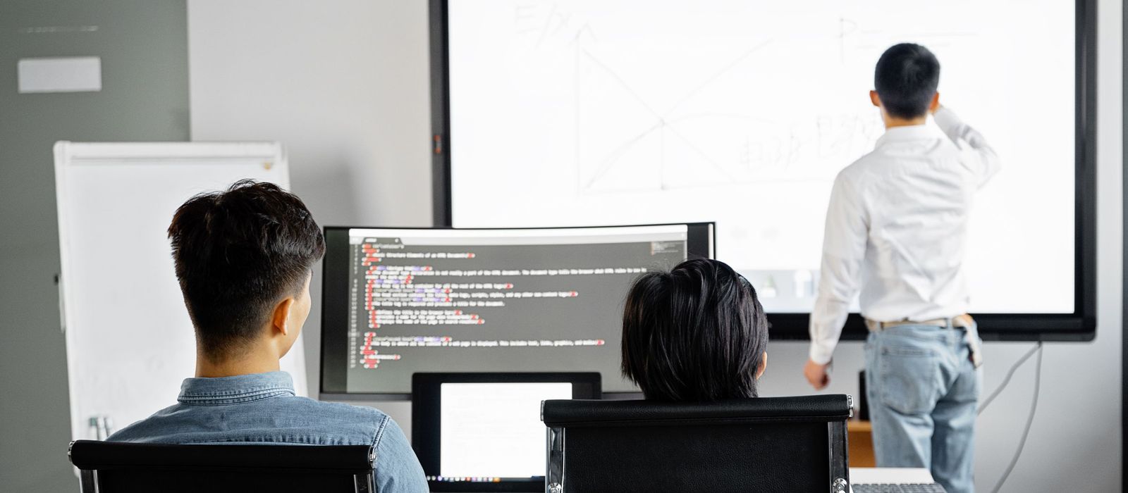 A training room seen from behind: two participants at their own screens while a trainer works through a diagram on the large wall screen